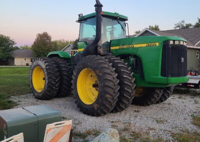 A John Deere 9300 tractor parked on gravel, with large yellow tires and a distinct green body, situated near traffic cones and a utility box in a residential area during sunset.