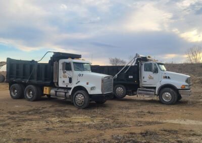Two dump trucks parked on a dirt lot, with one truck painted white and the other black, under a cloudy sky.