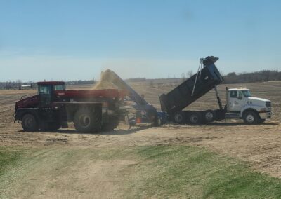A red agricultural tractor unloading material into a blue conveyor system, which is then transferring it into a white dump truck on a field under a clear blue sky.
