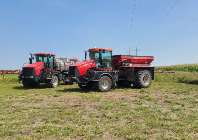 Two red agricultural tractors in a field, one equipped with a sprayer attachment, with power lines and a cornfield in the background.