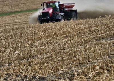A red agricultural tractor is working in a field, spreading fertilizer over a harvested corn crop. Dust is being kicked up from the ground, with power lines visible in the background. This image showcases modern farming practices essential for crop management.