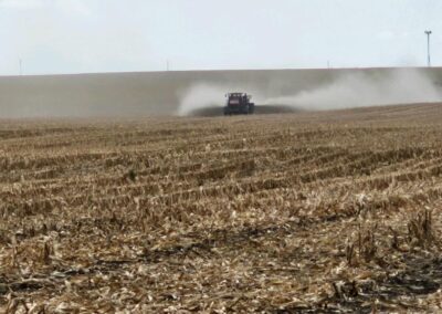 A tractor spraying fertilizer on a cornfield under a cloudy sky, with a dust trail visible behind it.
