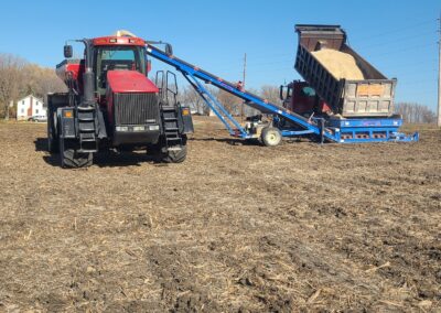 A red tractor is positioned in a field, connected to a blue grain cart that is tipping its contents. The background features a clear blue sky and a distant farmhouse.