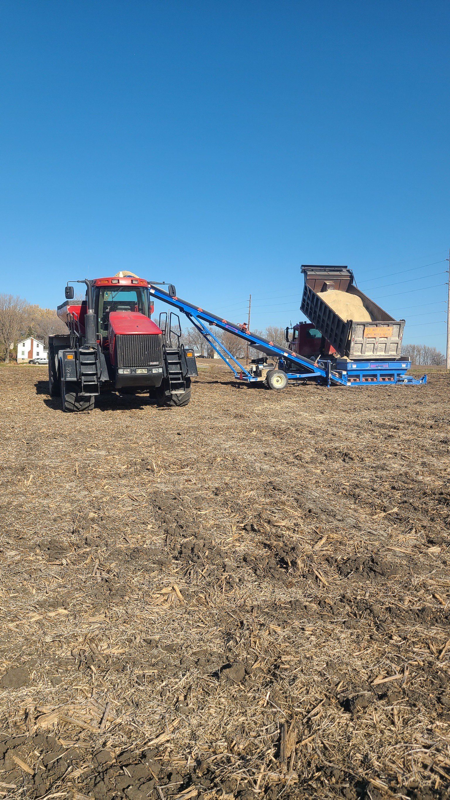 A red tractor positioned in a field with a blue conveyor system transferring material from a dump trailer, showcasing agricultural equipment used for farming operations at Millennial Ag Solutions.