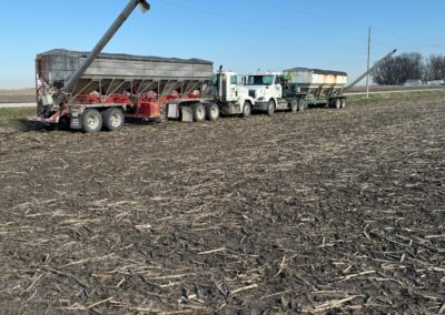 A red agricultural trailer with a conveyor, parked beside two large trucks in a field with dry soil and blue sky.
