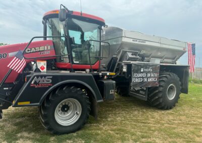 A red Case IH Titan tractor parked on grass, featuring a New Leader fertilizer spreader, an American flag attached, and a sign reading "Founded in Faith, Forged in America.