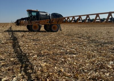A large agricultural sprayer is positioned in a harvested cornfield under a clear blue sky, with crop residue scattered across the ground. The sprayer's boom extends outwards, casting a shadow on the field.
