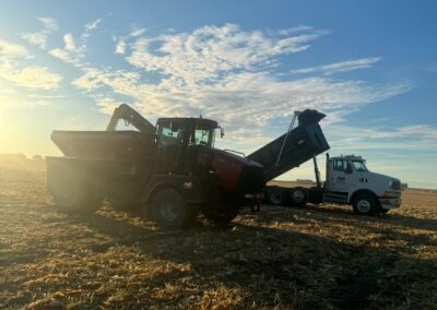 A tractor unloading corn into a truck on a field during sunset, with a clear blue sky and wispy clouds in the background.