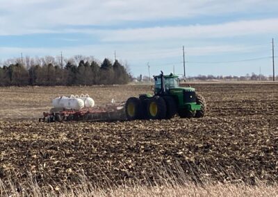 A green tractor with a tiller is working in a plowed field, pulling tanks for fertilizer or other agricultural substances in the background.