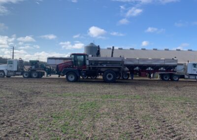 A red agricultural truck and a white semi-truck are parked on a field, with a large storage facility in the background under a partly cloudy sky.