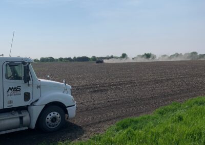 A white truck parked on the edge of a field with freshly tilled soil, while a tractor is working in the distance, creating a dust cloud under a clear blue sky. Lush green grass is visible in the foreground.
