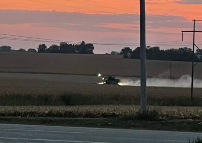 A combine harvester working in a field at dusk, with a colorful sunset in the background and power lines visible.