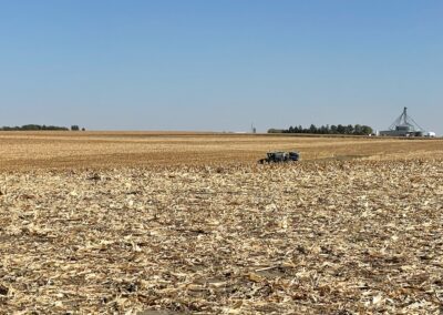 A vast agricultural field with harvested corn stubble, a tractor working in the distance, and a grain storage facility visible under a clear blue sky.