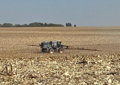 A tractor equipped with a sprayer is navigating through a vast, harvested cornfield under a clear sky.