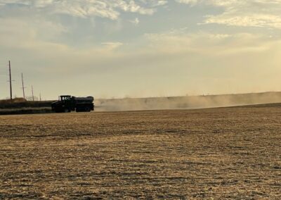A farming vehicle is working in a vast, dry field under a cloudy sky, creating dust as it moves.